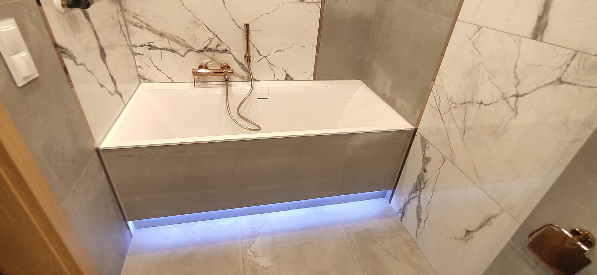 Nowoczesna łazienka z białą wanną obudowaną szarymi płytkami, podświetlenie LED u dołu, marmurowe płytki na ścianach i złota armatura.