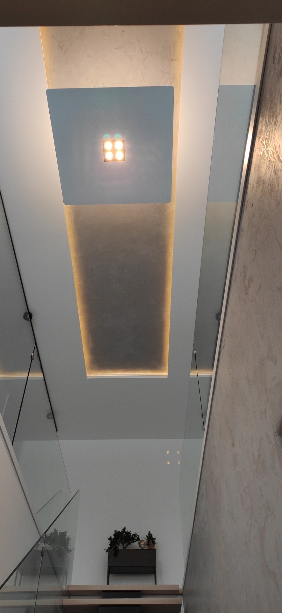 Nowoczesne wnętrze klatki schodowej z oświetleniem LED w suficie, szklane balustrady i dekoracyjna donica z roślinami na podeście.