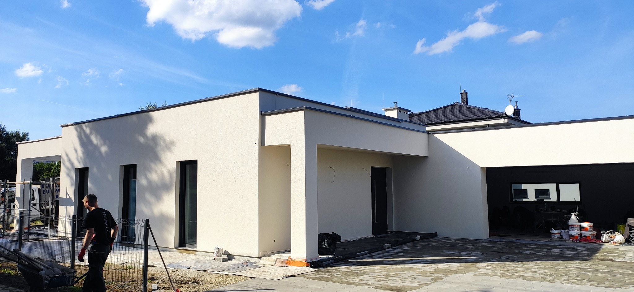 Nowoczesny dom jednorodzinny z jasną elewacją i czarnymi ramami okiennymi, widoczny pracownik budowlany i materiały budowlane na podjeździe.