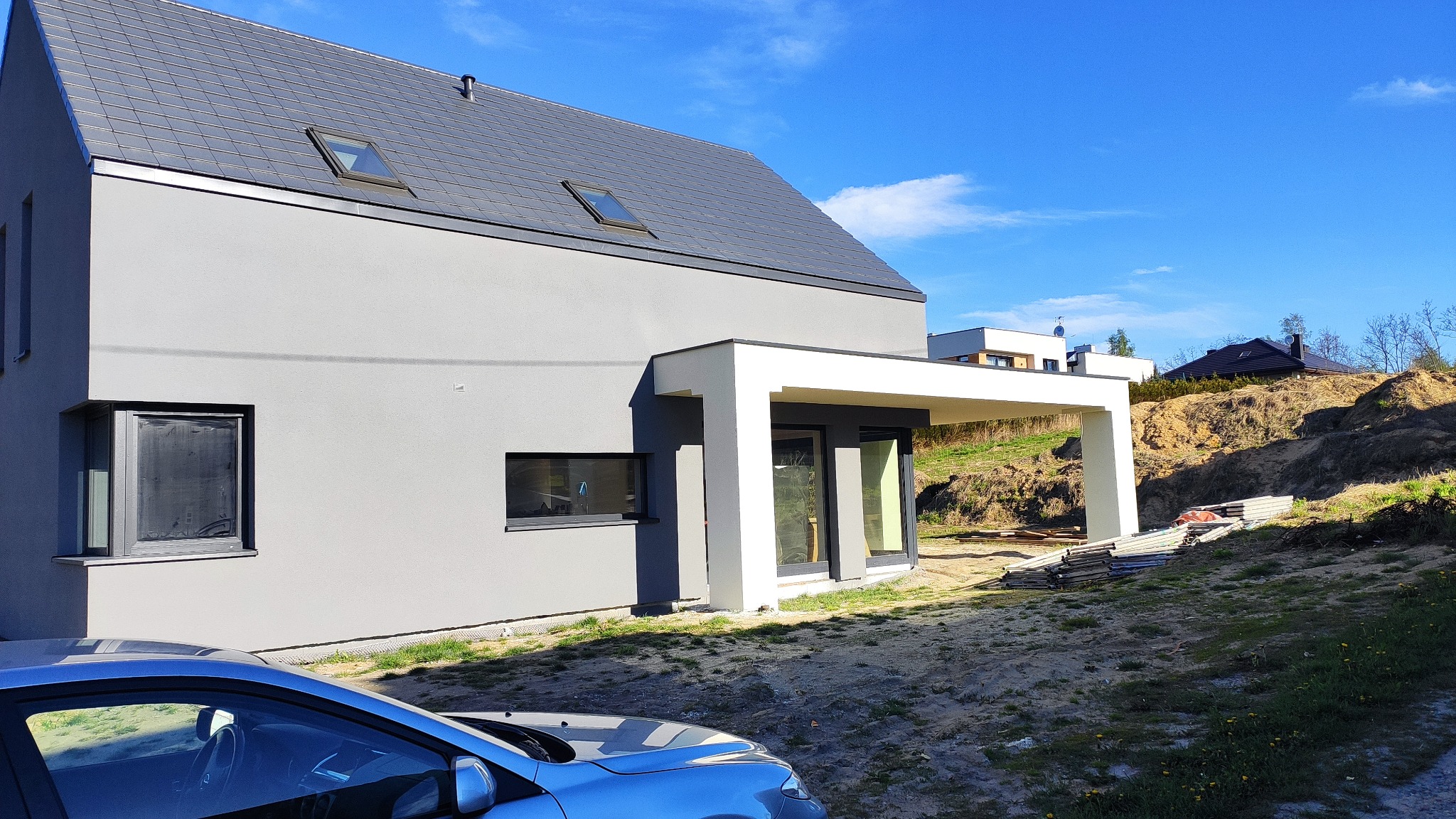 Nowoczesny dom jednorodzinny z szarą elewacją i grafitową dachówką, widoczny fragment niebieskiego samochodu na pierwszym planie, teren wokół domu częściowo zagospodarowany.