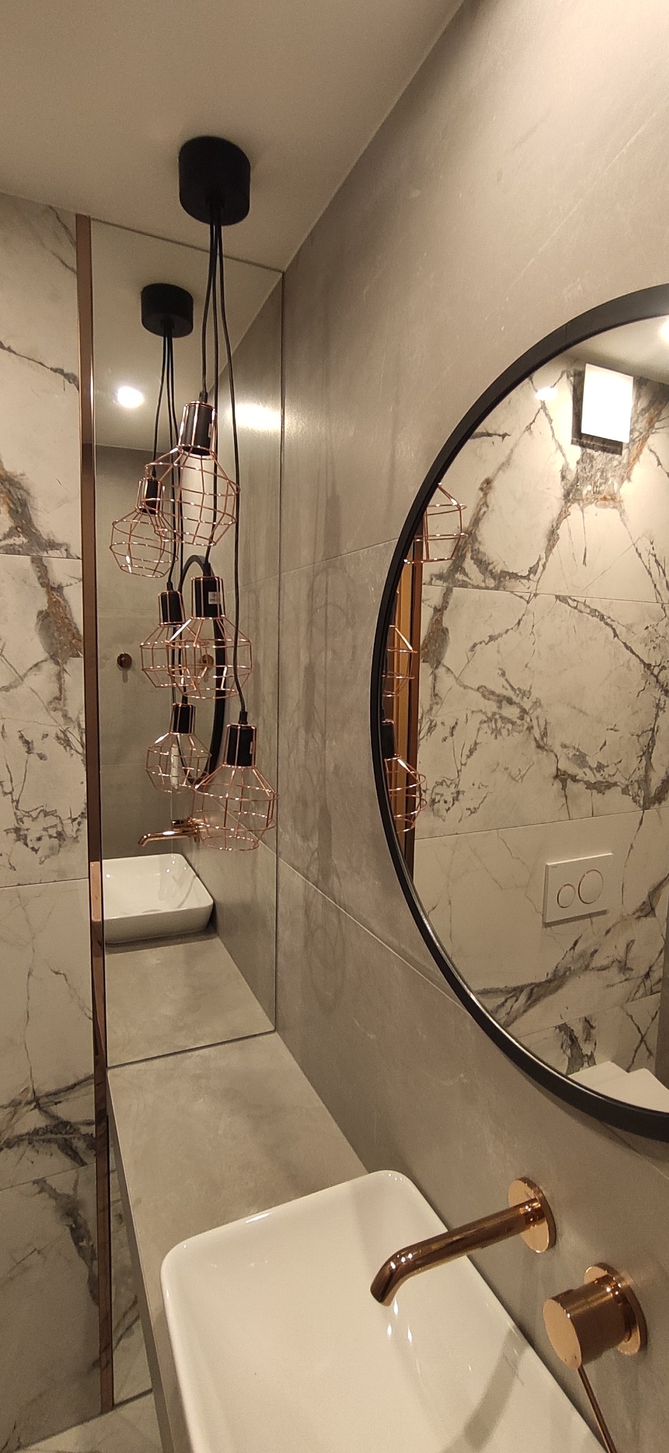 Nowoczesna łazienka z marmurowymi płytkami, okrągłym lustrem i wiszącymi lampami w stylu industrialnym z miedzianymi akcentami.