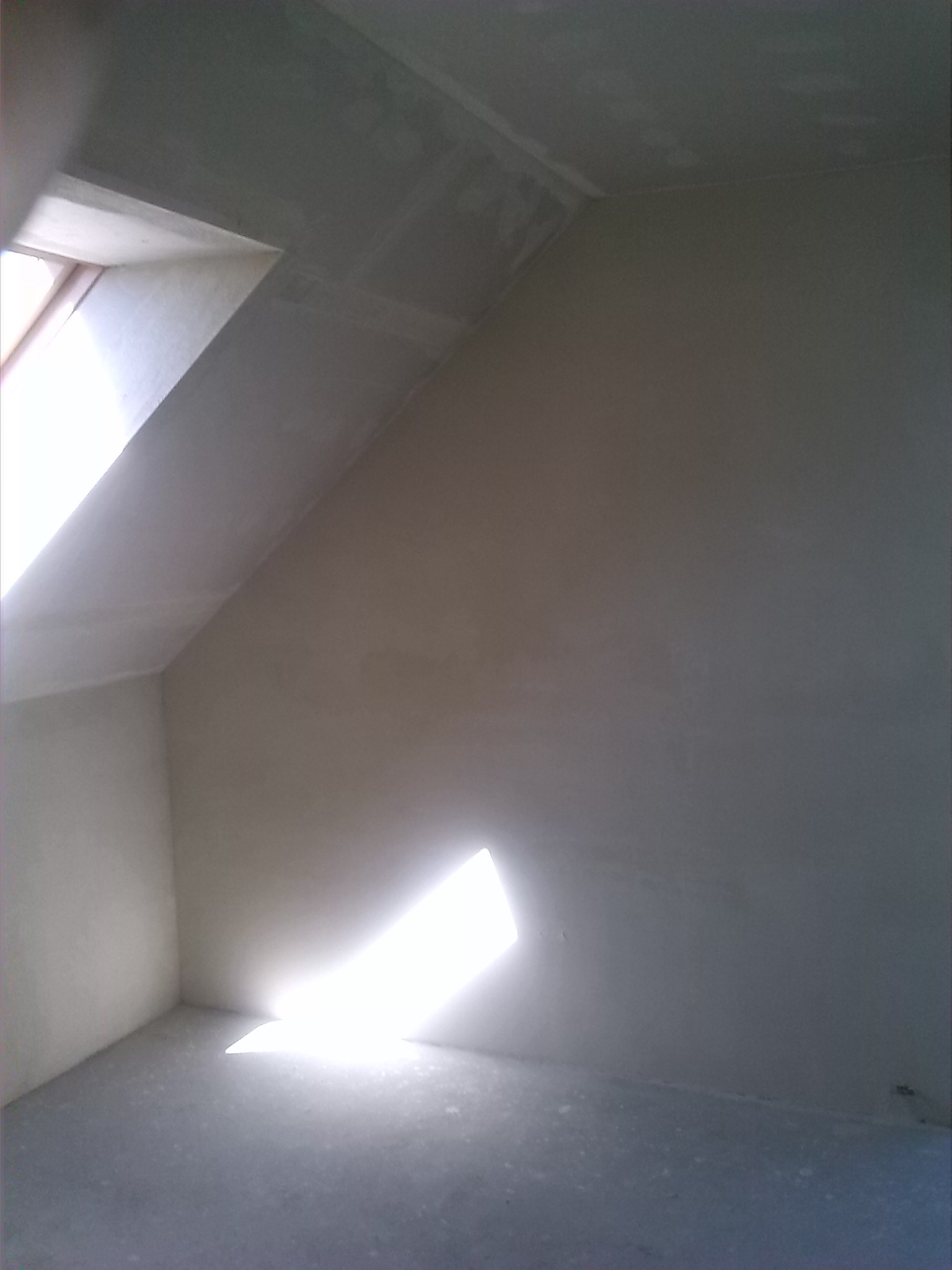 Poddasze ze świeżo nałożoną gładzią szpachlową, widoczne okno dachowe i plama światła na podłodze.