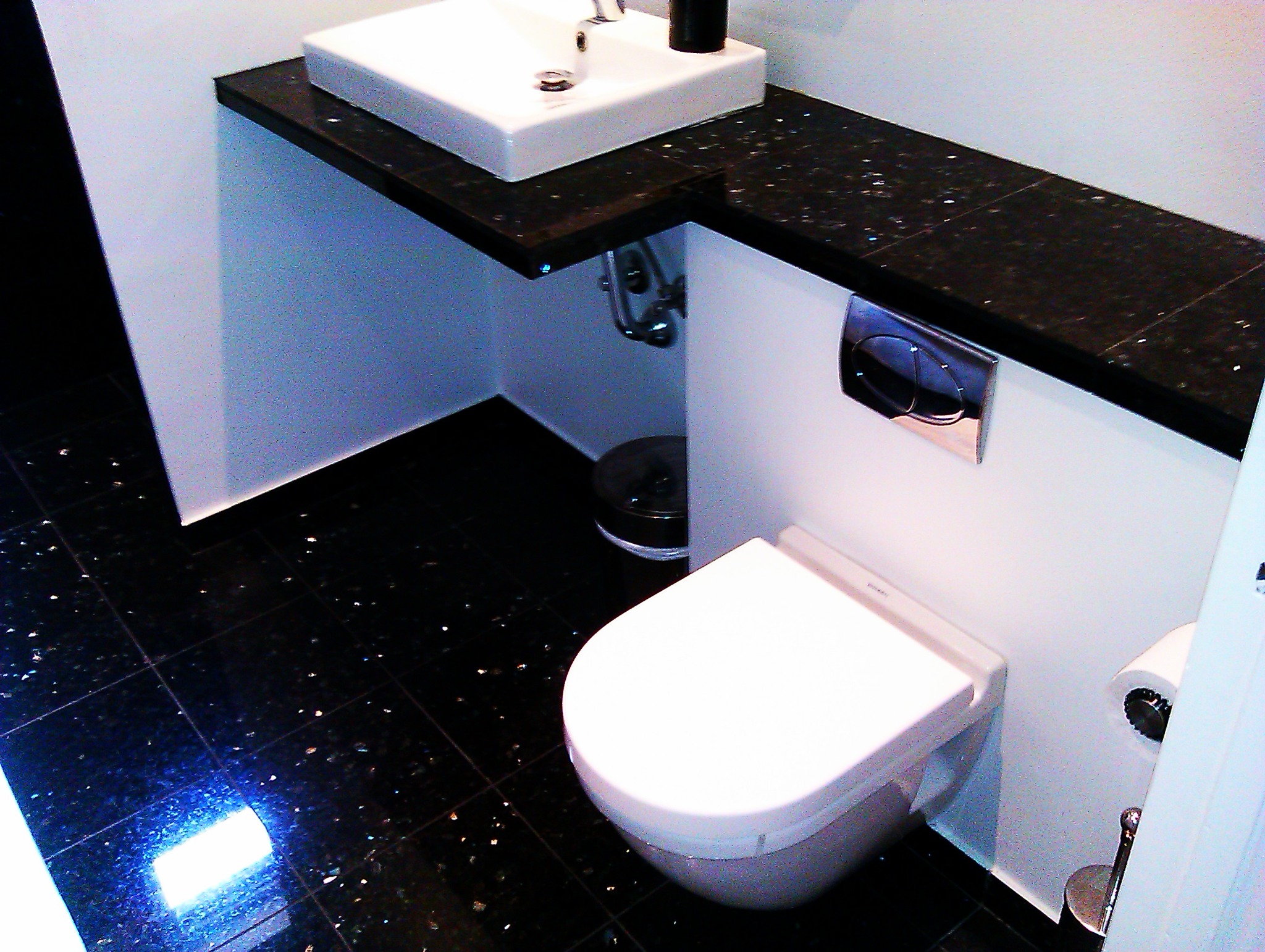Nowoczesna łazienka z czarną, błyszczącą posadzką i blatem, białą umywalką i podwieszaną toaletą z chromowanym przyciskiem spłukiwania, widoczne elementy instalacji pod umywalką.