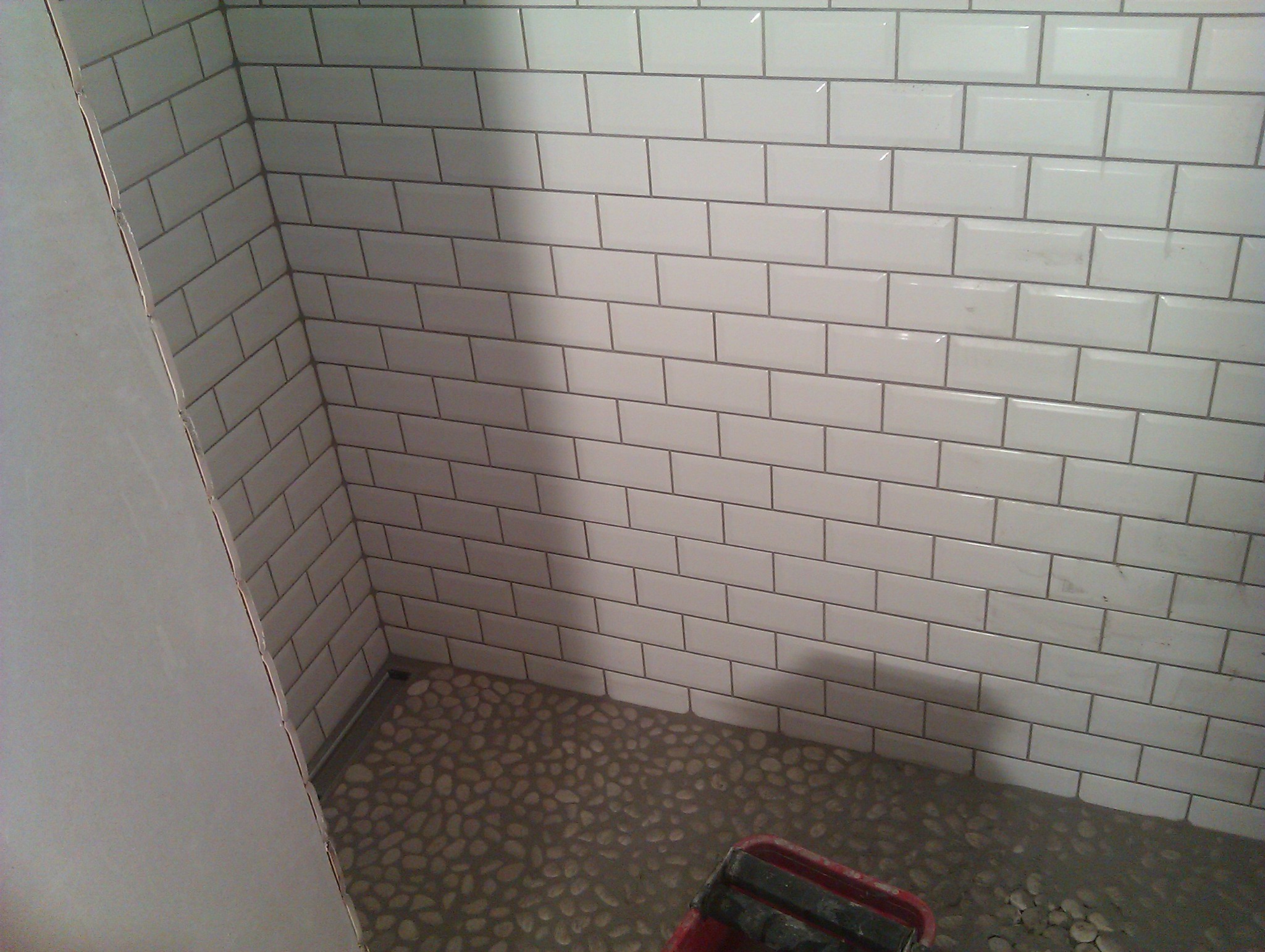 Świeżo ułożone białe, błyszczące płytki w kształcie cegiełek na ścianie w rogu łazienki, z podłogą wyłożoną drobnymi, okrągłymi kamieniami i widocznym fragmentem czerwonego wiadra glazurniczego...