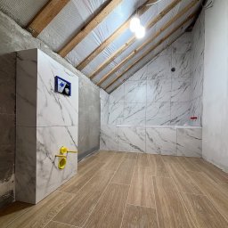 Quick Refit Daniel Rychlewski - Wykończona łazienka na poddaszu w Warszawie. Marmurowe płytki ścienne i drewnopodobna podłoga, zabudowa WC z żółtymi elementami. Widoczne drewniane belki stropowe.