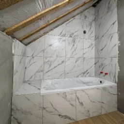 Wykończona łazienka na poddaszu ze skośnym sufitem. Białe płytki imitujące marmur okalają wannę. Widoczne drewniane elementy konstrukcji dachu.