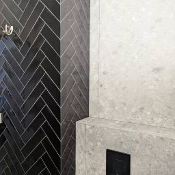 Quick Refit Daniel Rychlewski - Czarne, błyszczące płytki ułożone w jodełkę na ścianie łazienki, kontrastujące z jasną, marmurkową ścianą z czarnym przyciskiem spłuczki.