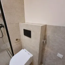 Nowoczesna łazienka z beżowymi płytkami imitującymi kamień. Wisząca toaleta z czarnym przyciskiem spłukującym. Widoczny fragment kabiny prysznicowej z czarnymi profilami.