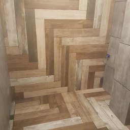 Quick Refit Daniel Rychlewski - Ułożona w jodełkę podłoga z płytek imitujących drewno, z widocznym odpływem liniowym przy ścianie i fragmentem ściany wyłożonej płytkami w odcieniach szarości.