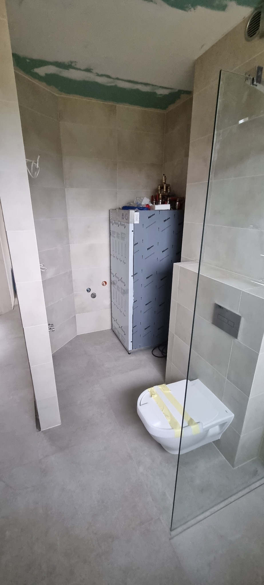 Łazienka w trakcie remontu, widoczna zabudowa geberit z folią ochronną, płytki na ścianach i podłodze, sufit z widocznymi śladami prac wykończeniowych, zamontowana kabina prysznicowa.