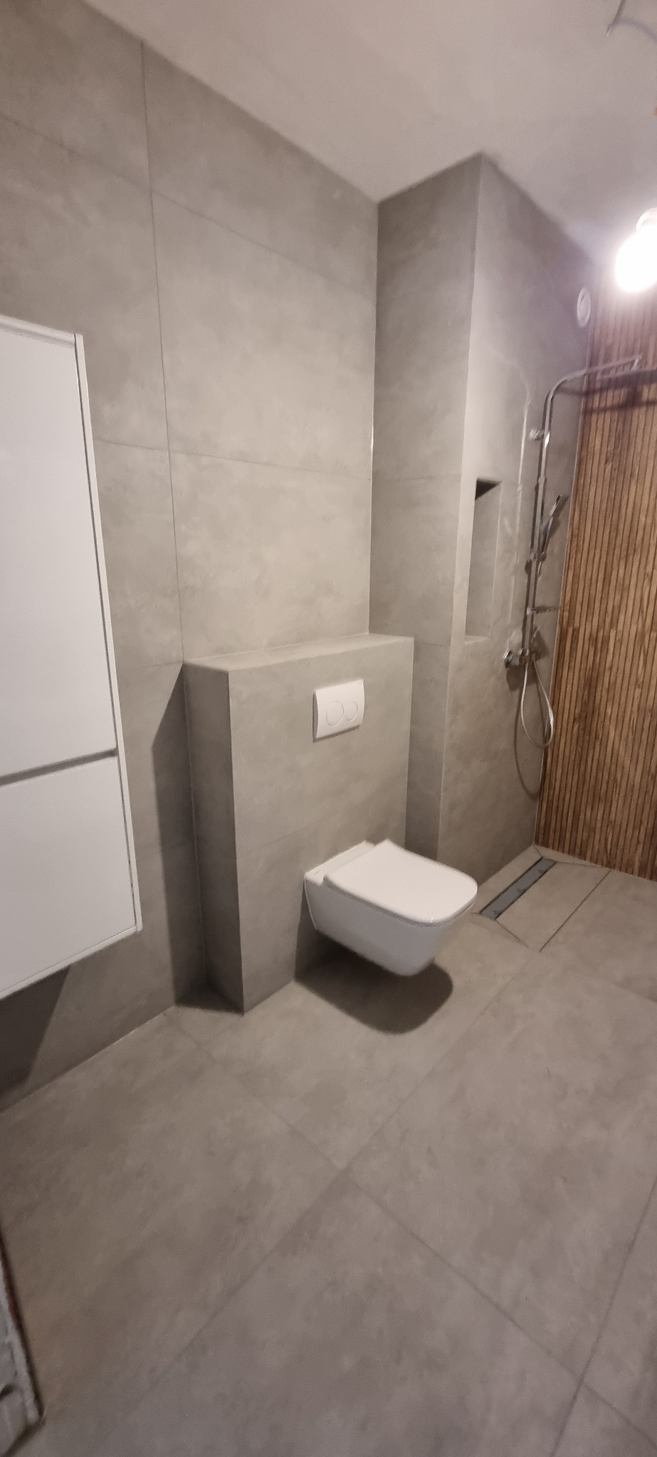 Nowoczesna łazienka z szarą płytką, podwieszaną toaletą z białym przyciskiem, kabiną prysznicową z drewnianymi panelami i odpływem liniowym.