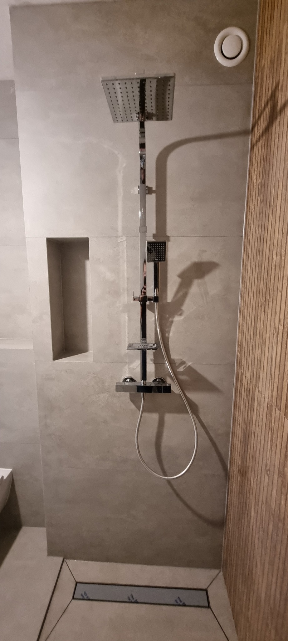 Nowoczesna kabina prysznicowa z deszczownicą i odpływem liniowym, wykończona płytkami w odcieniach szarości, z drewnianym akcentem na ścianie po prawej stronie i wnęką w ścianie po lewej.