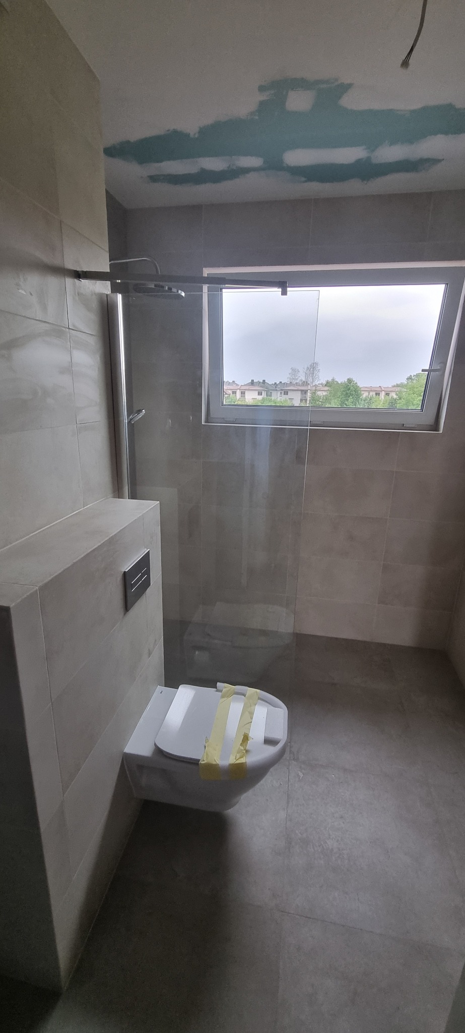 Nowoczesna łazienka w trakcie remontu: szare płytki, kabina prysznicowa ze szklaną ścianą, podwieszana toaleta zabezpieczona taśmą malarską, sufit z widocznymi śladami gładzi