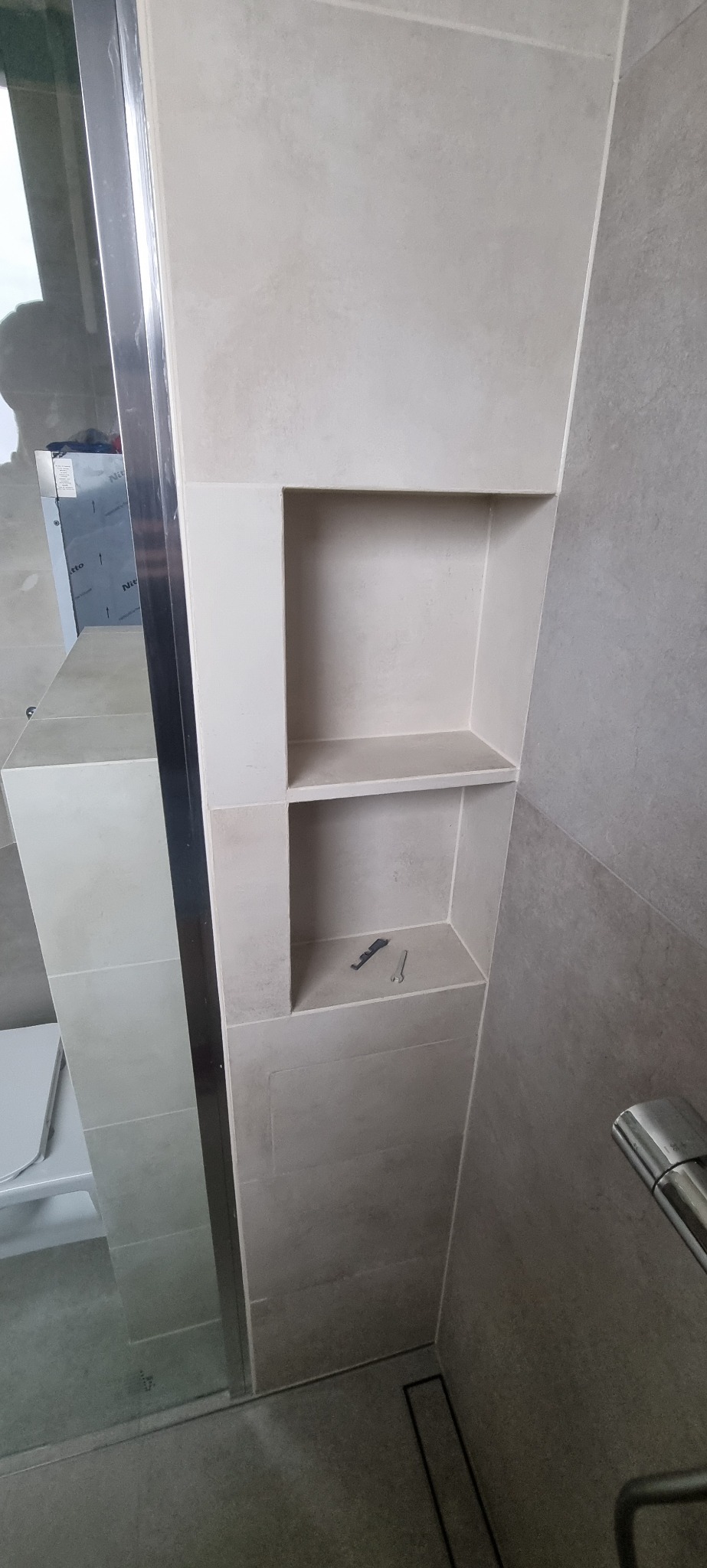 Świetlisty beżowy prysznic z wnęką wyłożoną płytkami, w której leżą narzędzia, widoczna szklana kabina i chromowana armatura.