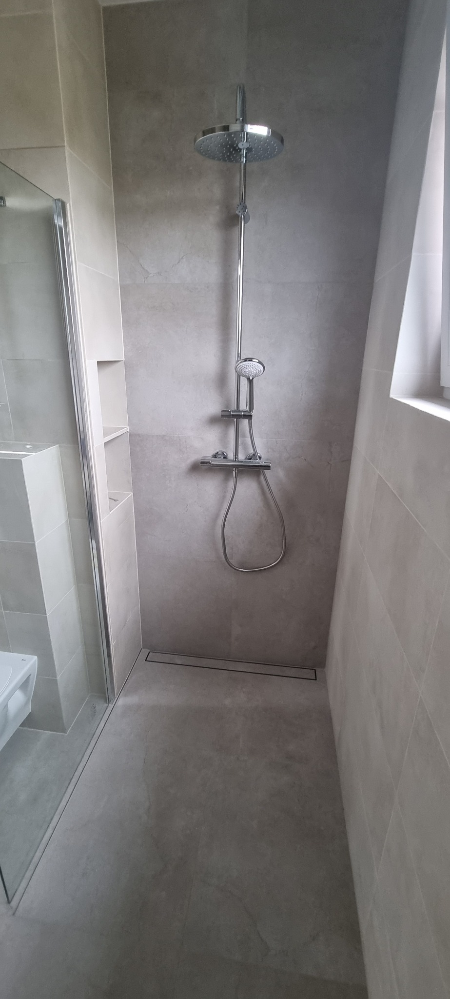 Nowoczesna kabina prysznicowa walk-in z deszczownicą, wykończona szarymi płytkami wielkoformatowymi, z odpływem liniowym i wnęką półkową.