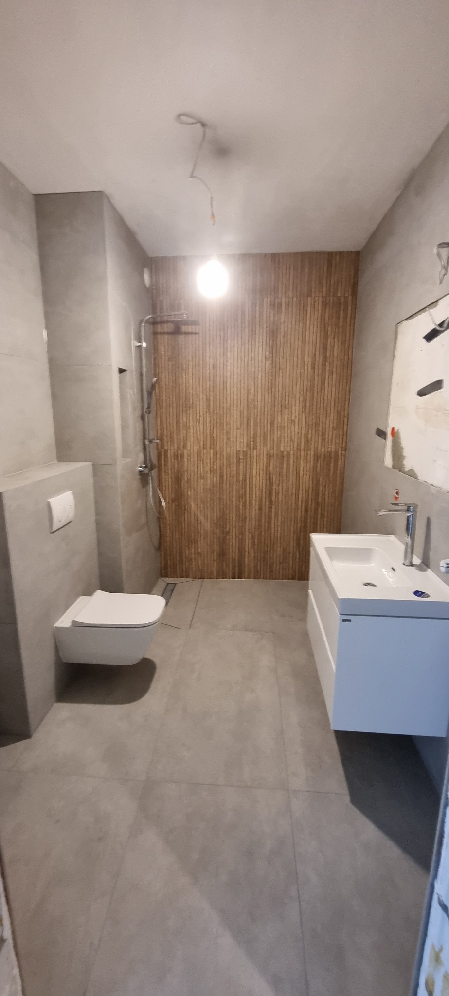 Nowoczesna łazienka z wiszącą toaletą, umywalką na szafce i prysznicem z drewnianymi panelami na ścianie, widoczne niedokończone prace wykończeniowe na ścianie.