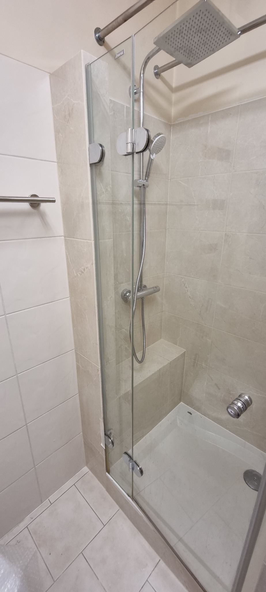 Nowoczesna kabina prysznicowa z jasnymi płytkami, szklanymi drzwiami z chromowanymi zawiasami i deszczownicą.