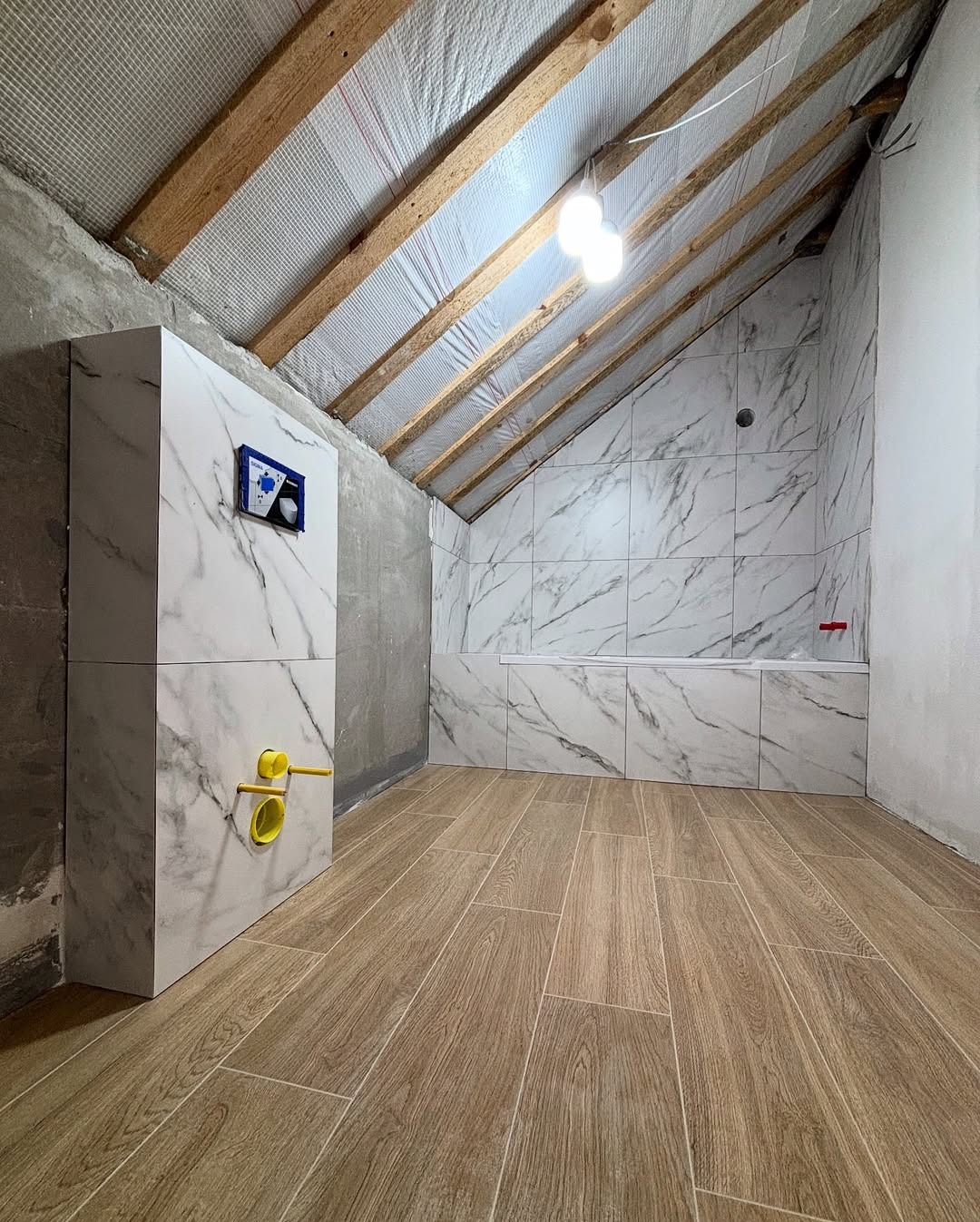 Wykończona łazienka na poddaszu w Warszawie. Marmurowe płytki ścienne i drewnopodobna podłoga, zabudowa WC z żółtymi elementami. Widoczne drewniane belki stropowe.