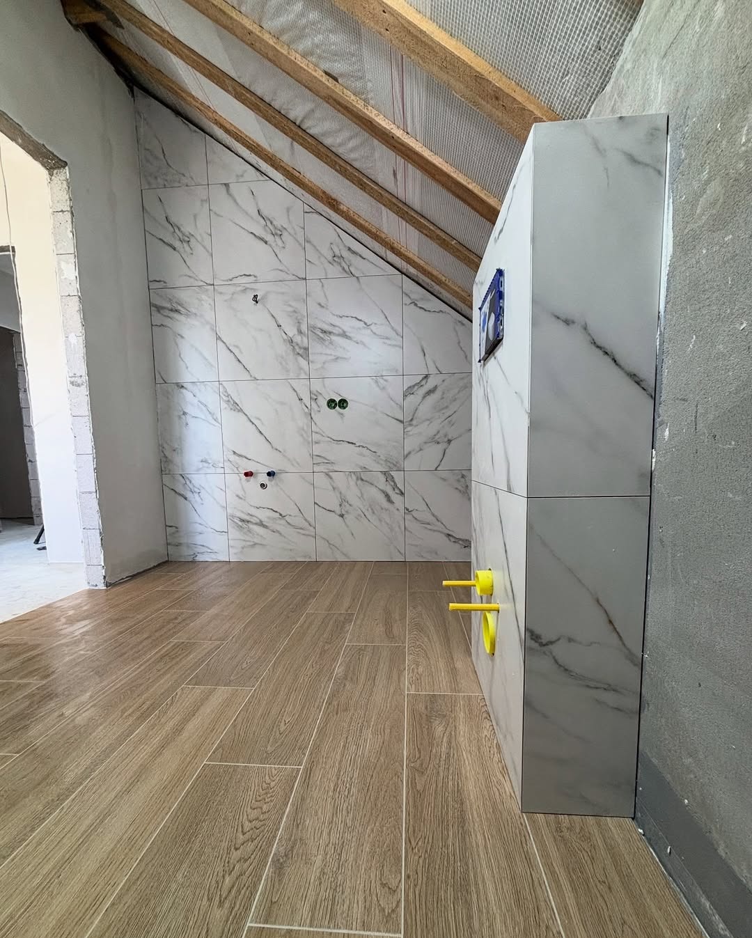 Łazienka w trakcie wykańczania: marmurowe płytki na ścianach pod skosem, drewnopodobna podłoga, zabudowa stelaża WC z żółtymi elementami instalacji.