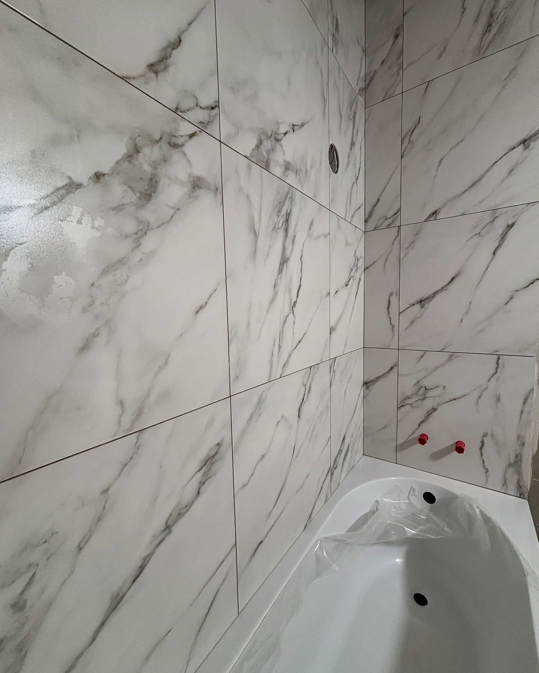 Białe marmurowe płytki ścienne w łazience, widoczna wanna zabezpieczona folią. Minimalistyczny design, czyste linie, nowoczesne wykończenie.