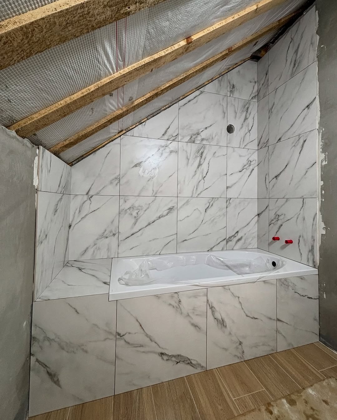 Wykończona łazienka na poddaszu ze skośnym sufitem. Białe płytki imitujące marmur okalają wannę. Widoczne drewniane elementy konstrukcji dachu.