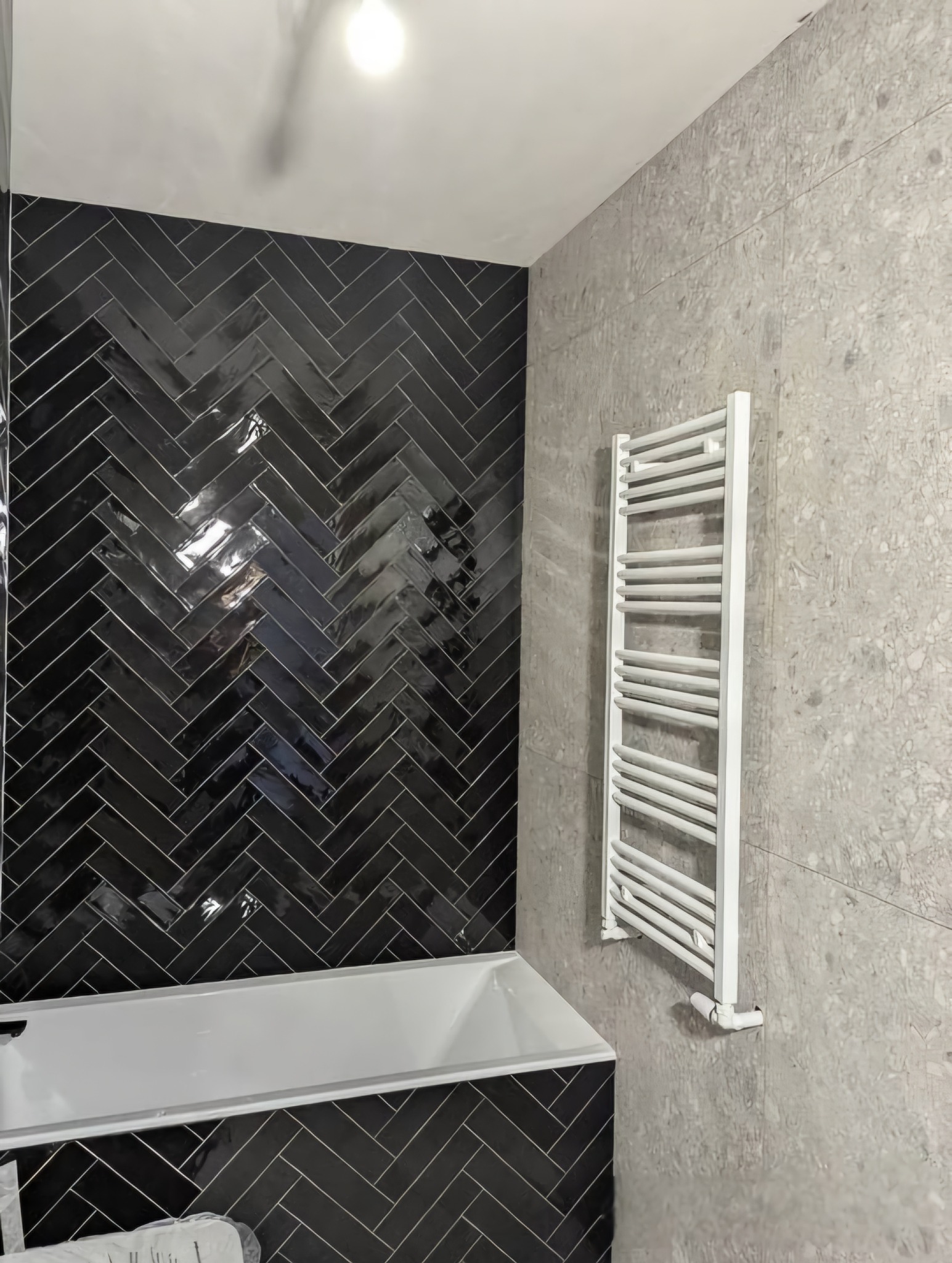 Łazienka po remoncie: czarna, błyszcząca glazura ułożona w jodełkę nad wanną, szara ściana z grzejnikiem drabinkowym. Nowoczesny design.