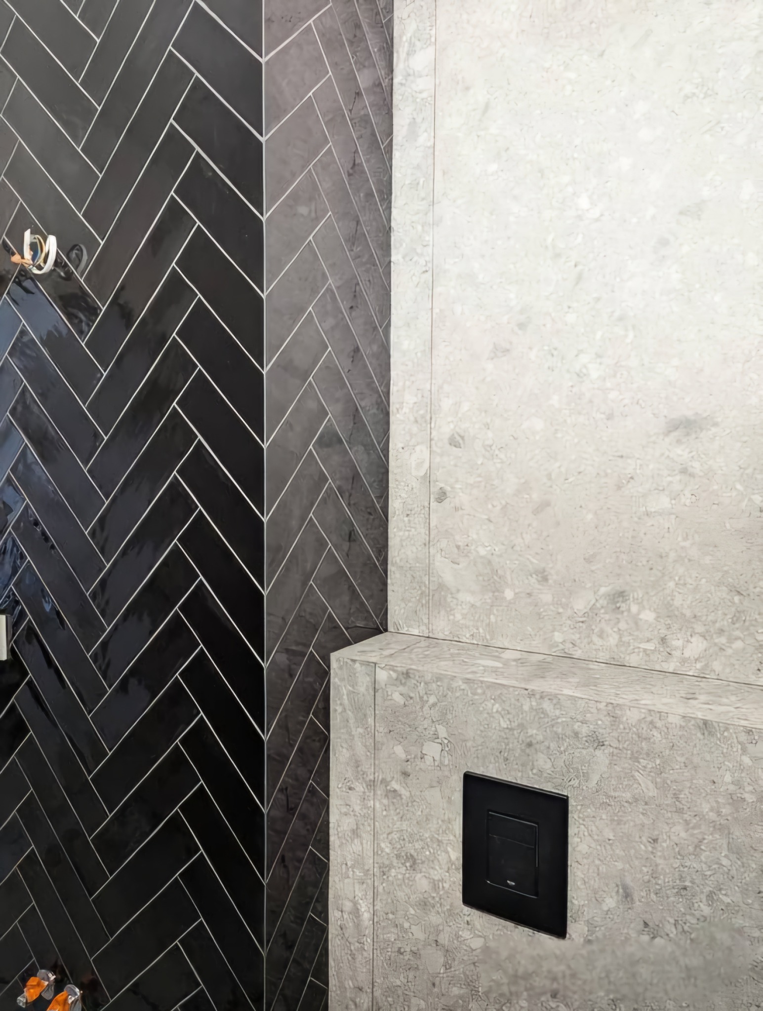 Czarne, błyszczące płytki ułożone w jodełkę na ścianie łazienki, kontrastujące z jasną, marmurkową ścianą z czarnym przyciskiem spłuczki.