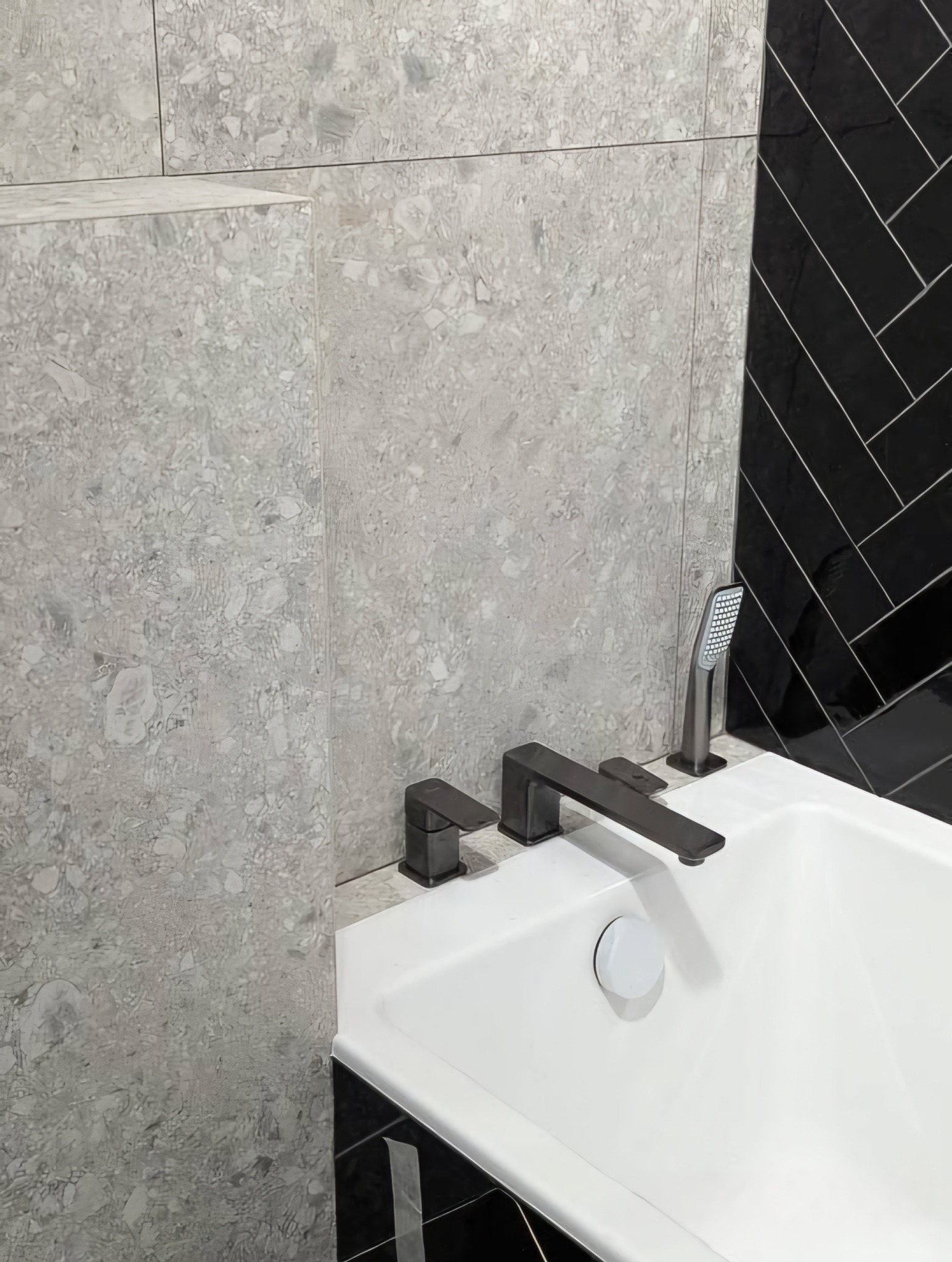 Biała wanna z ciemną armaturą na tle ściany z szarej mozaiki i czarnych płytek ułożonych w jodełkę. Nowoczesne wykończenie łazienki.