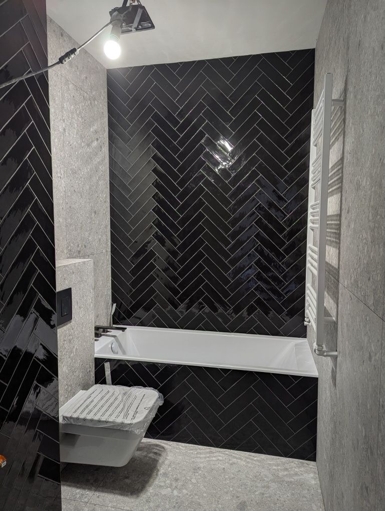 Ciemna łazienka w jodełkę: czarne, błyszczące płytki na ścianie za wanną, szare płytki na ścianie bocznej i podłodze. Biała wanna i grzejnik.
