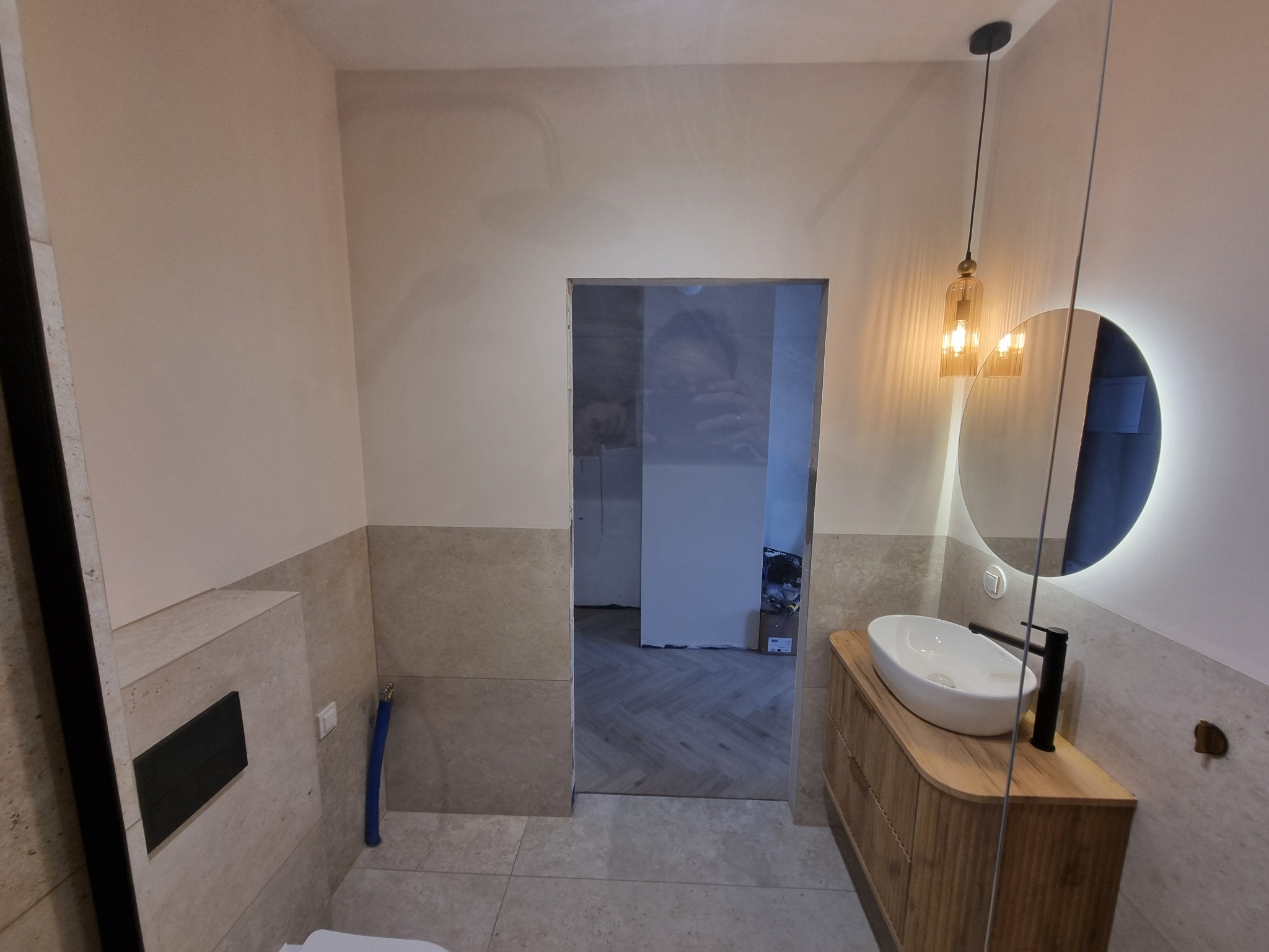 Wykończona łazienka z umywalką na drewnianej szafce, okrągłym lustrem z podświetleniem i wiszącą lampą. Na ścianach płytki do połowy wysokości i gładź. Widok na sąsiednie pomieszczenie.