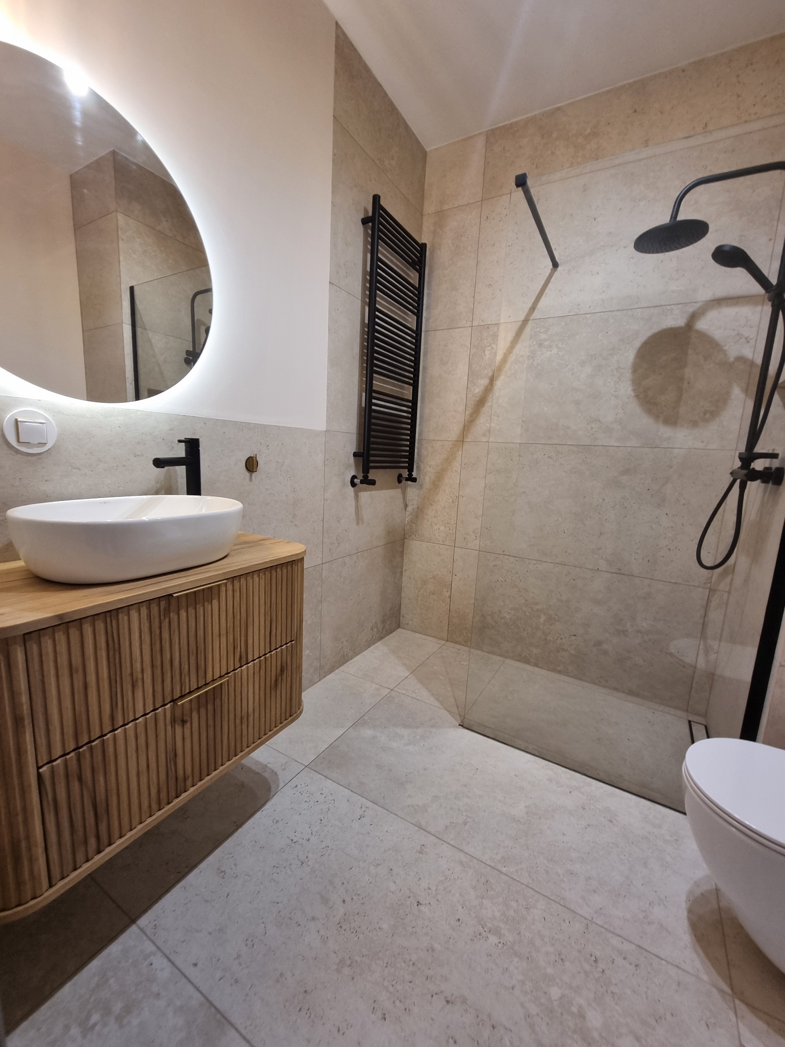 Nowoczesna łazienka z umywalką nablatową na drewnianej szafce, okrągłe lustro z podświetleniem LED i czarna armatura. Beżowe płytki na ścianach i podłodze.