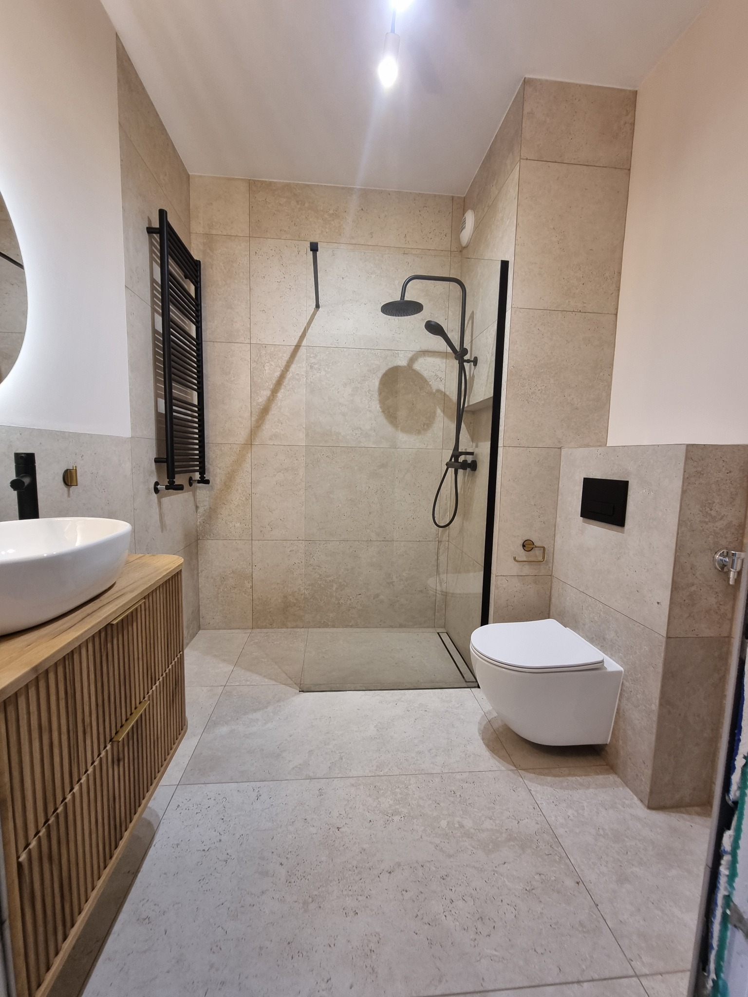 Jasna łazienka z beżowymi płytkami, czarną armaturą i drewnianą szafką pod umywalką. Kabina prysznicowa typu walk-in z czarnymi profilami. Umywalka nablatowa.