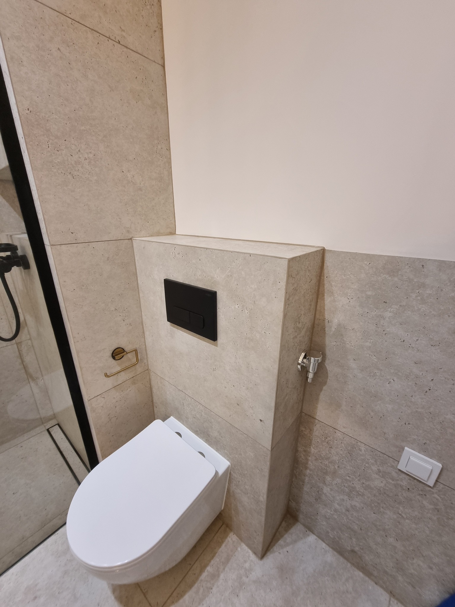 Nowoczesna łazienka z beżowymi płytkami imitującymi kamień. Wisząca toaleta z czarnym przyciskiem spłukującym. Widoczny fragment kabiny prysznicowej z czarnymi profilami.