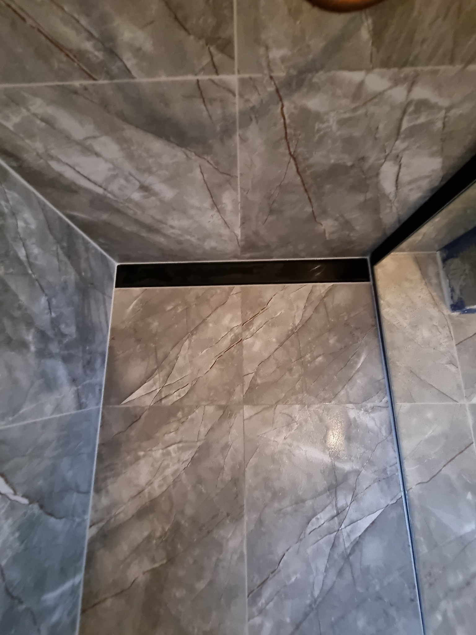 Szare płytki imitujące marmur z ciemną fugą w narożniku kabiny prysznicowej. Widok z góry, podkreślający wzór i precyzję ułożenia płytek.