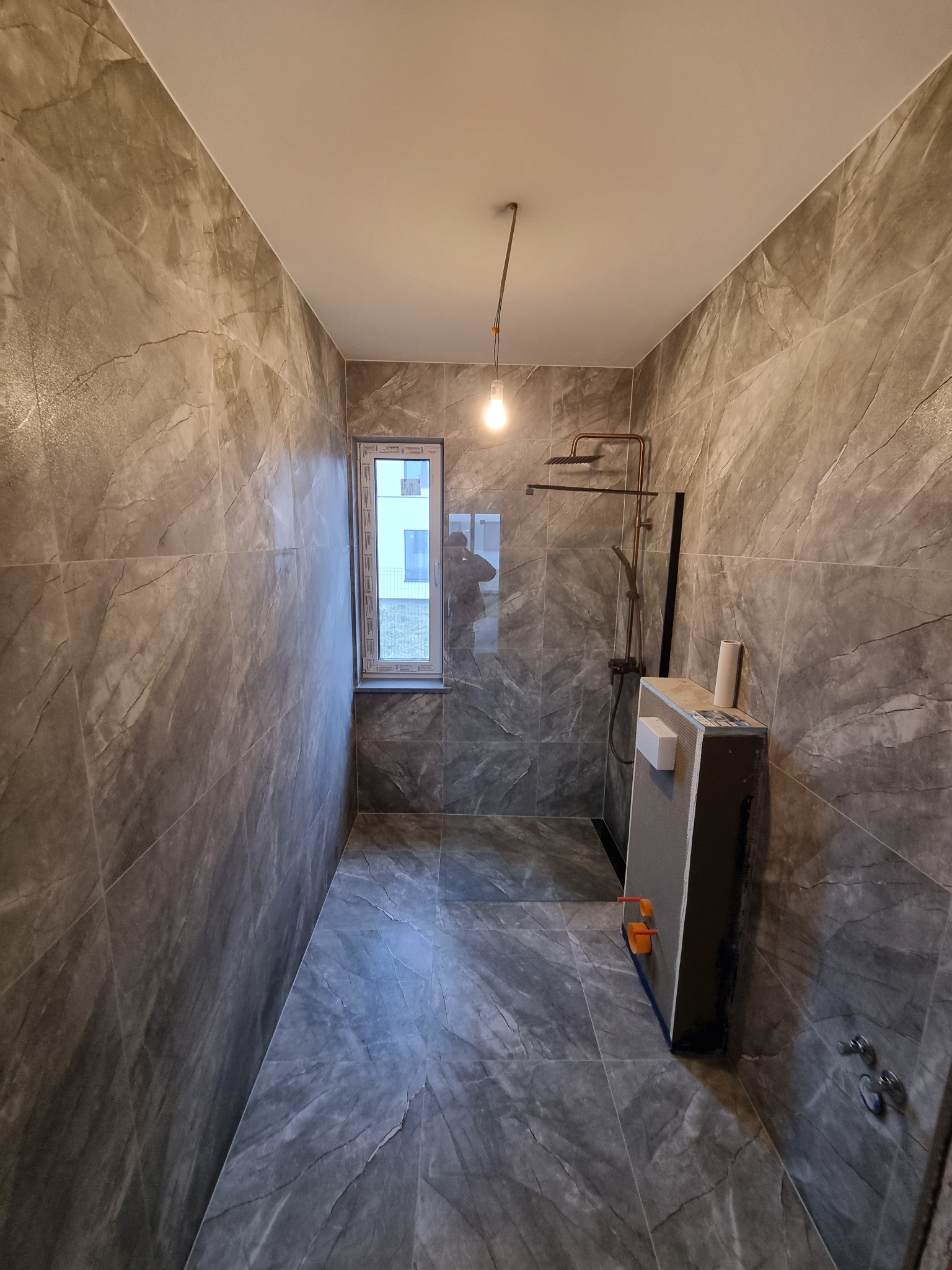 Wykończona łazienka z szarymi płytkami imitującymi marmur, widoczna kabina prysznicowa z miedzianą armaturą i okno. Minimalistyczny design, surowy styl.