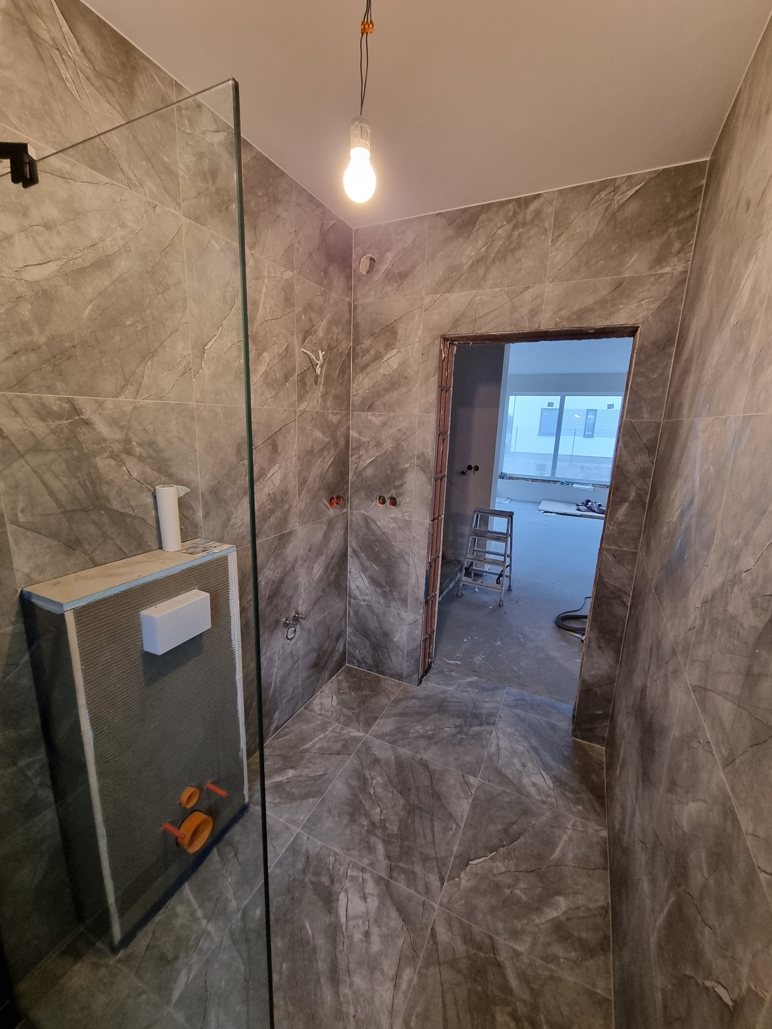 Wykończona łazienka z szarymi płytkami imitującymi marmur, widoczna szklana kabina prysznicowa i stelaż podtynkowy WC. W tle drabina i widok na inne pomieszczenie.