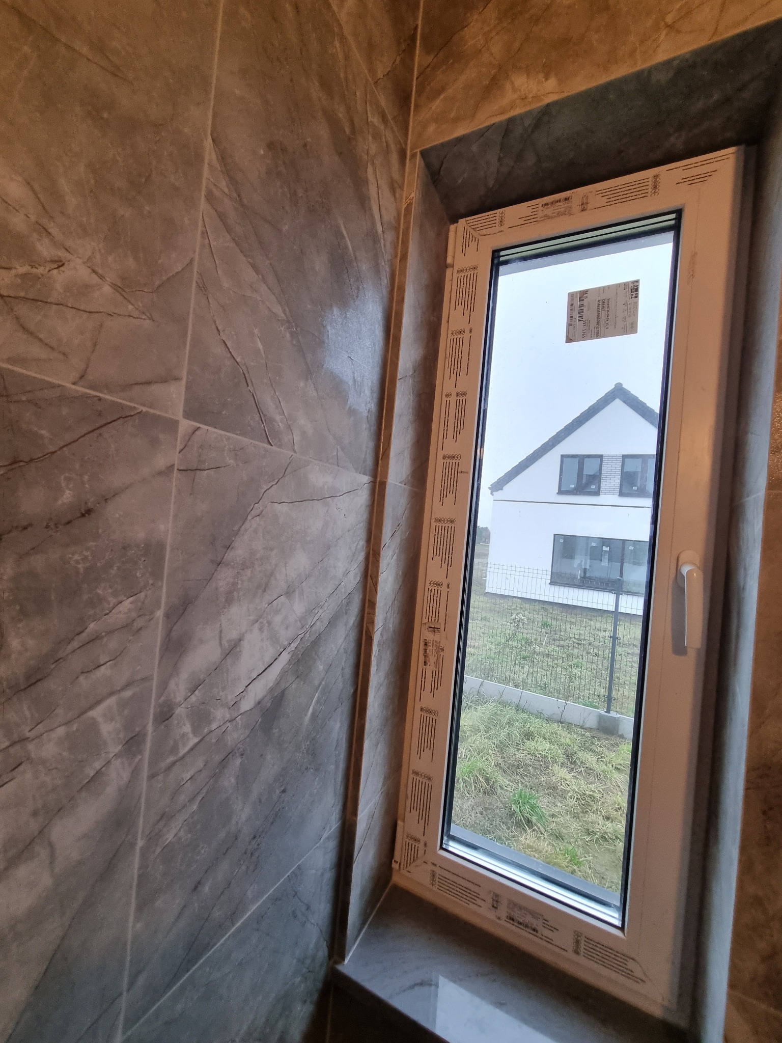 Szara, marmurkowa okładzina ścienna w łazience z oknem PCV, przez które widać dom. Wykończenie w nowoczesnym stylu, z dbałością o detale.