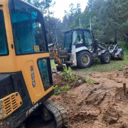 Dwa traktory: żółty minikoparka JCB 8025 ZTS z naklejką ostrzegawczą na szybie i biało-czarny Hidromek Alpha na terenie budowy z błotem i śladami opon.