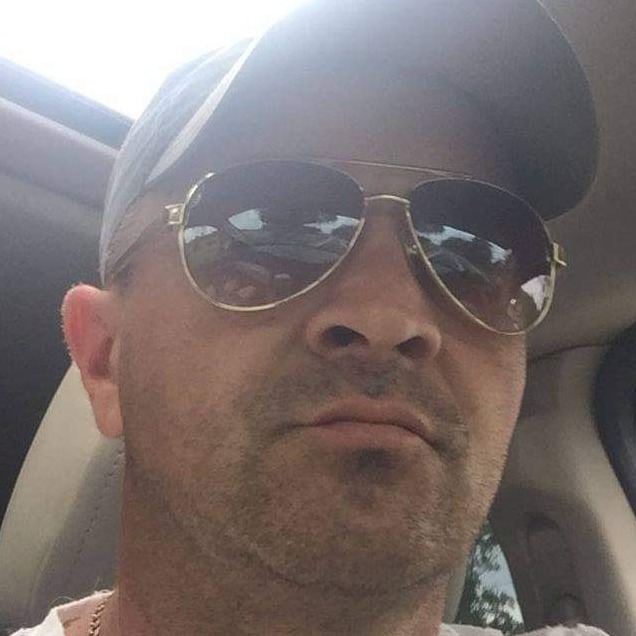 Portret mężczyzny w okularach przeciwsłonecznych i czapce z daszkiem, siedzącego w samochodzie z otwartym szyberdachem.