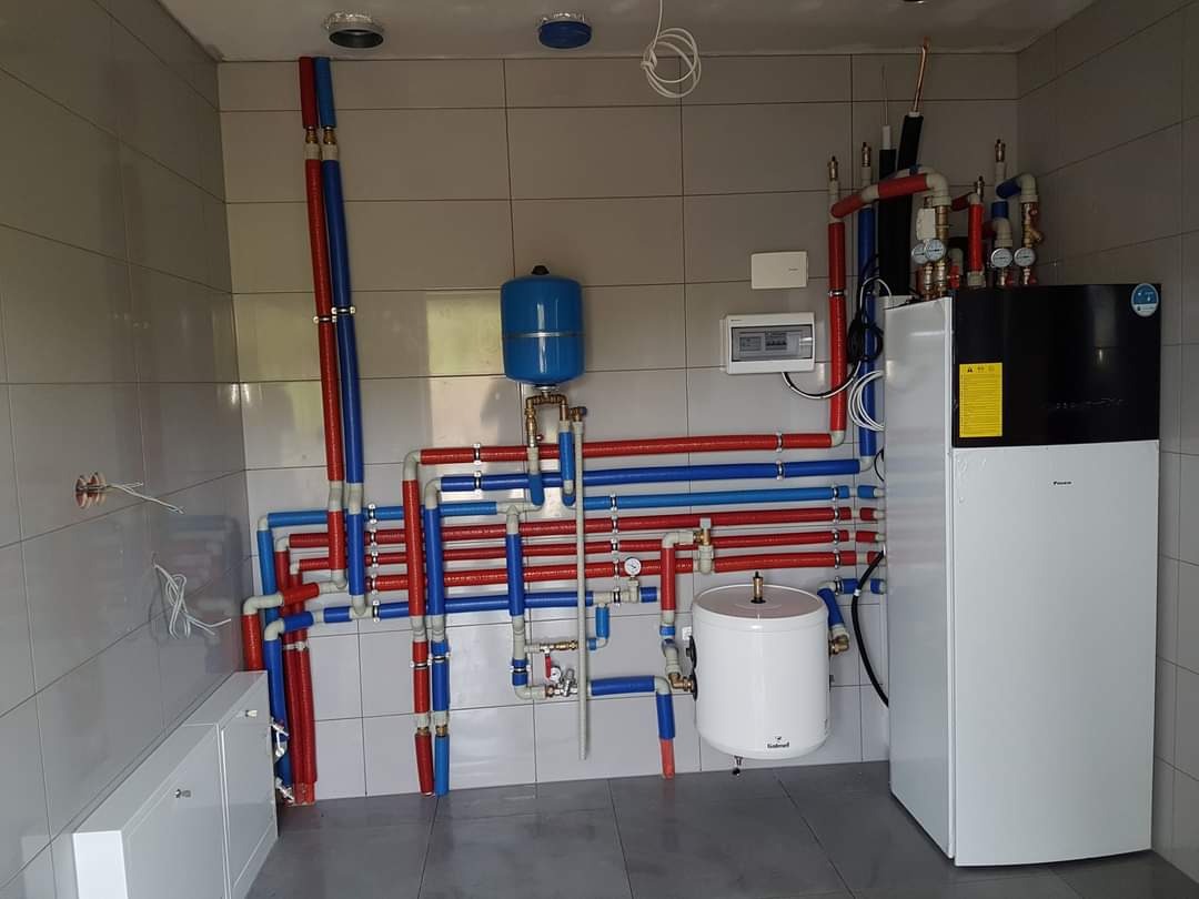 Kotłownia z piecem CO, zasobnikiem ciepłej wody i rozbudowaną instalacją hydrauliczną w kolorach czerwonym i niebieskim, na tle białych płytek.