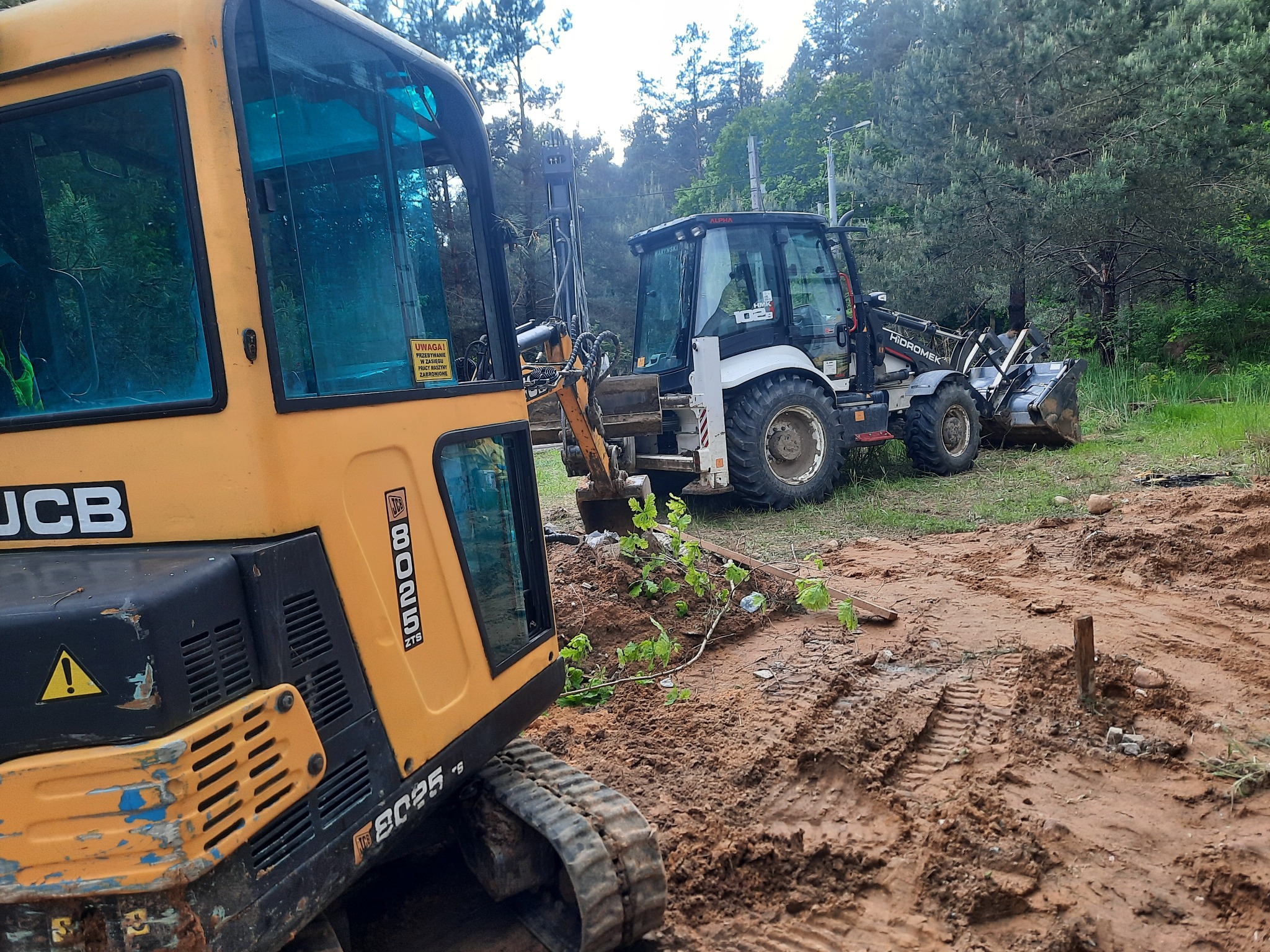 Dwa traktory: żółty minikoparka JCB 8025 ZTS z naklejką ostrzegawczą na szybie i biało-czarny Hidromek Alpha na terenie budowy z błotem i śladami opon.
