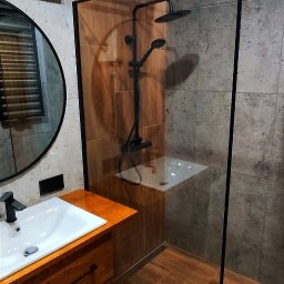 Finezja Remonty z Pomysłem - Nowoczesna łazienka z kabiną prysznicową typu walk-in z czarnymi okuciami, drewnianą szafką pod umywalką z blatem z litego drewna i okrągłym lustrem.