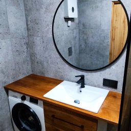 Finezja Remonty z Pomysłem - Nowoczesna łazienka z pralką zabudowaną pod drewnianym blatem umywalki, okrągłe lustro z czarną ramą, szare płytki na ścianach i brązowe płytki imitujące drewno na podłodze.