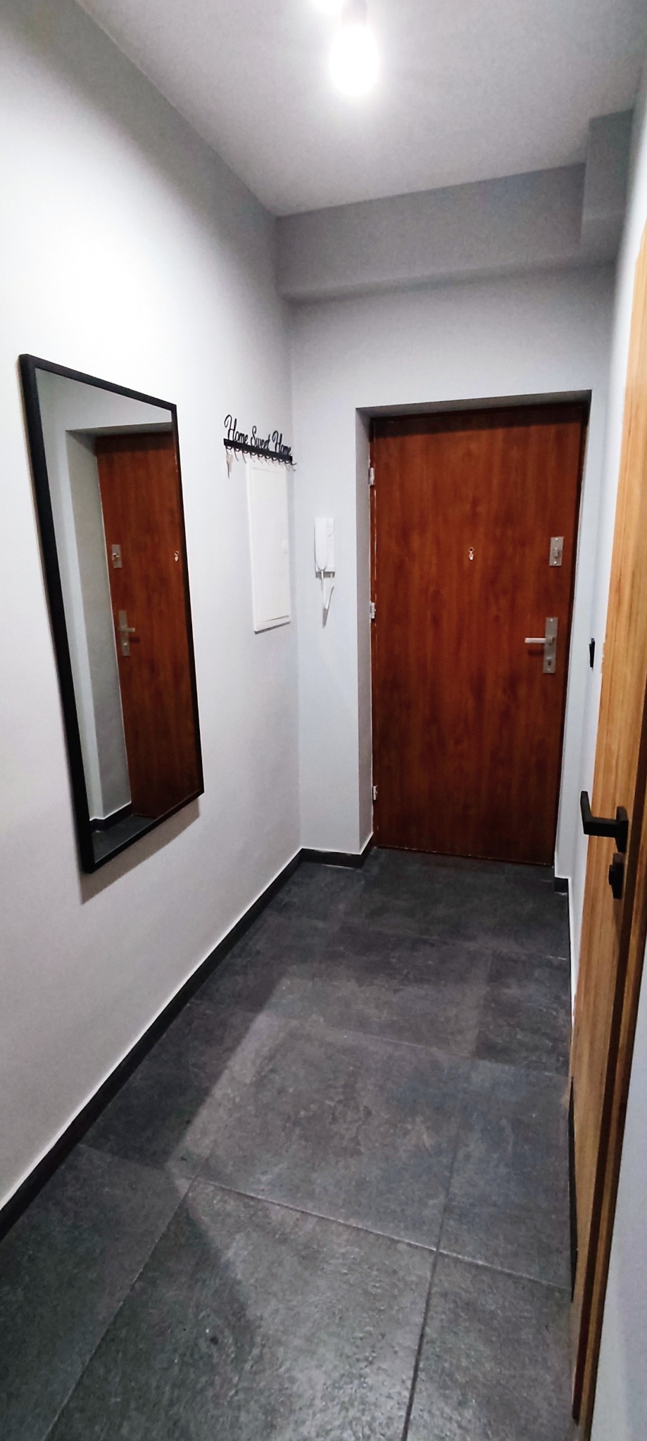 Szare płytki podłogowe w przedpokoju, drzwi wejściowe z brązowego drewna, lustro w czarnej ramie i wieszak na klucze z napisem 'Home Sweet Home' na ścianie.