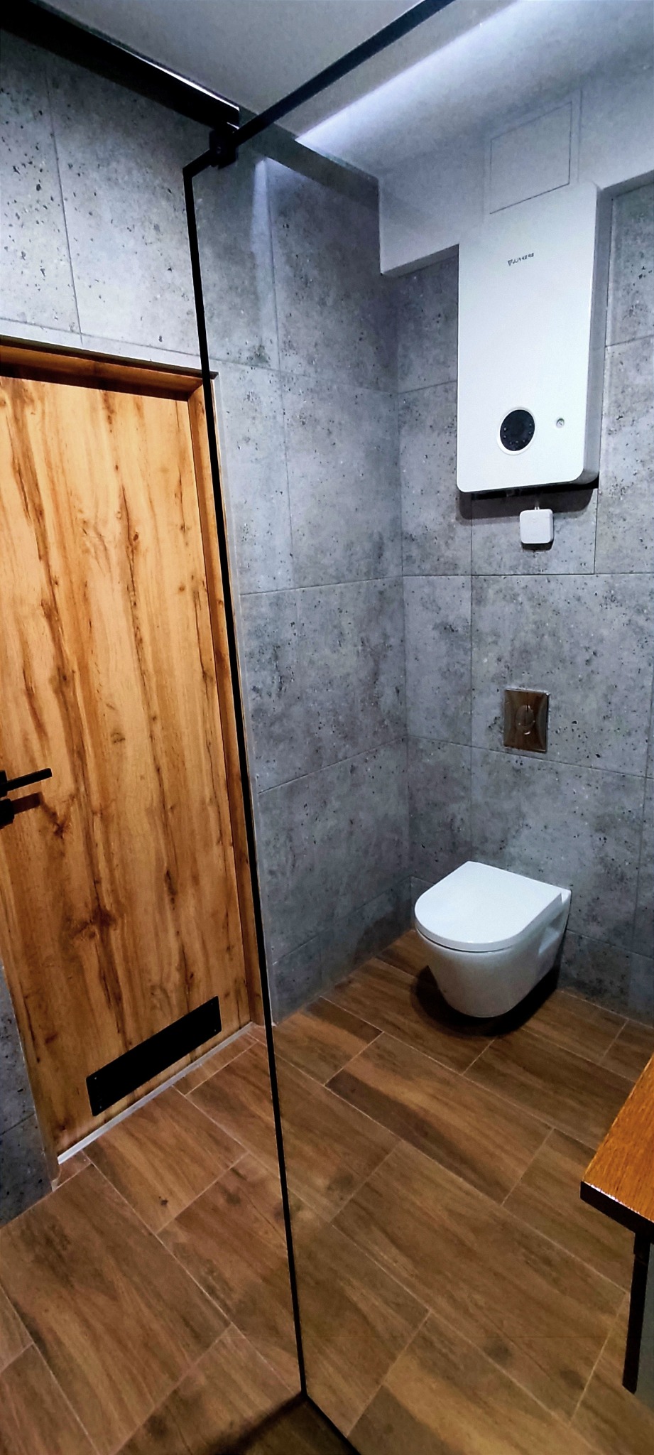 Nowoczesna łazienka z szarymi płytkami imitującymi kamień, drewnianą podłogą i drzwiami z czarną armaturą, wisząca toaleta i bojler Junkers na ścianie.