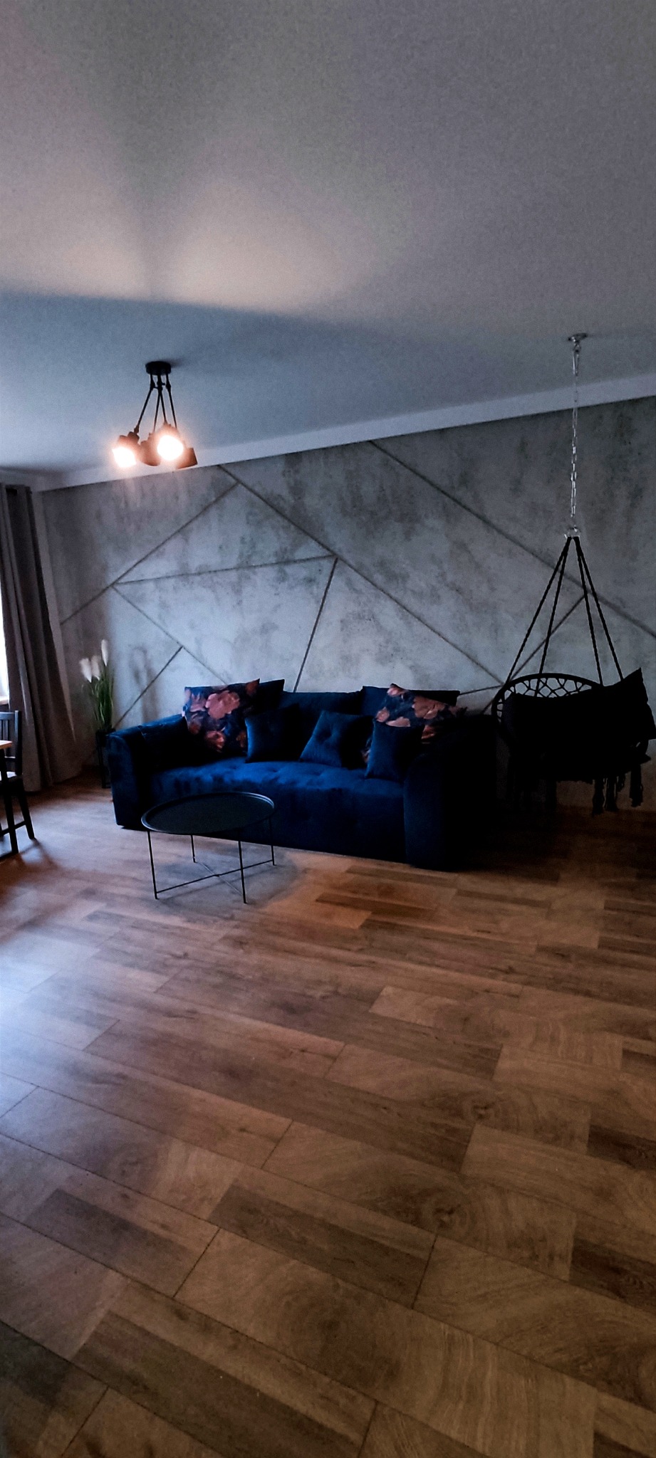 Nowoczesny salon z ciemnoniebieską sofą z poduszkami w kwiaty, ścianą z geometrycznym wzorem, wiszącym fotelem hamakowym i drewnianą podłogą.