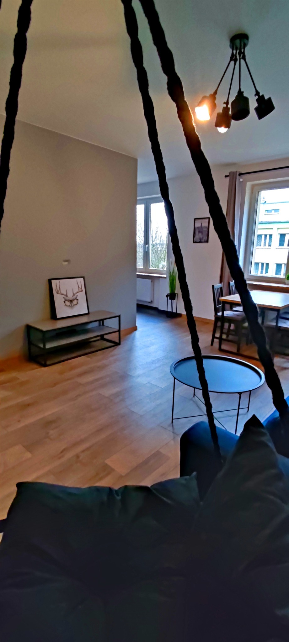 Jasne wnętrze mieszkania z drewnianą podłogą, widoczne z perspektywy wiszącego fotela, z czarnym stolikiem kawowym i minimalistycznym oświetleniem sufitowym.