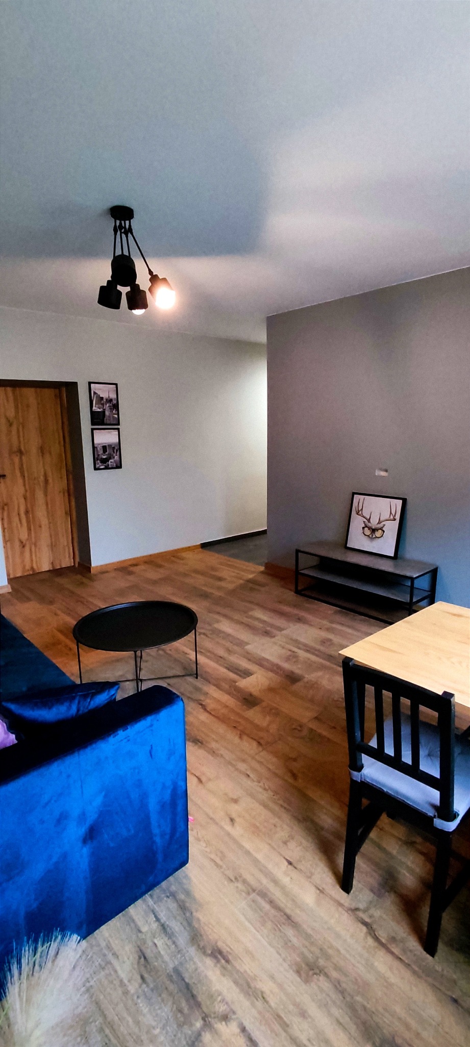 Nowoczesny salon z drewnianą podłogą, niebieską kanapą, czarnym stolikiem kawowym i lampą sufitową z trzema żarówkami, dekoracje ścienne w czarnych ramkach.