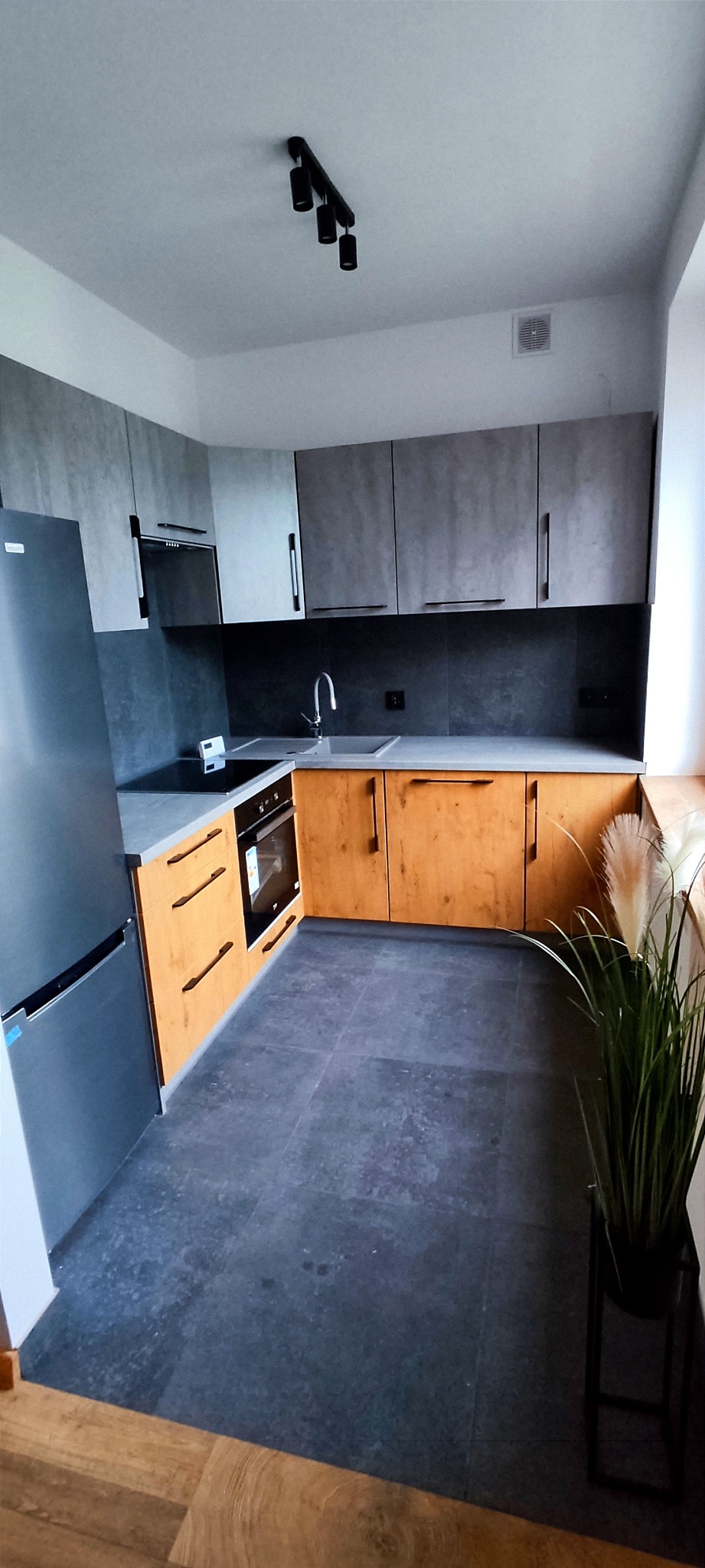 Nowoczesna kuchnia z szarymi górnymi szafkami i drewnopodobnymi dolnymi, czarnymi uchwytami, oraz czarną płytą indukcyjną i piekarnikiem; szara podłoga i roślina w czarnej donicy przy oknie.