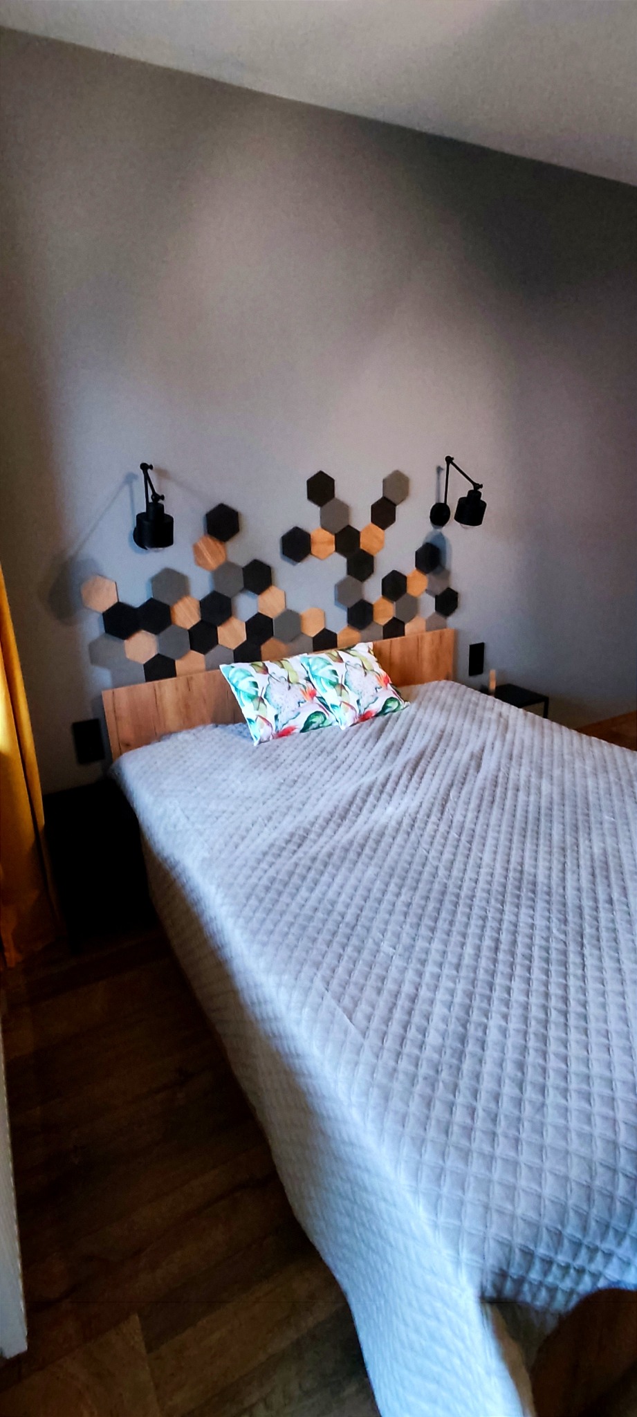 Sypialnia z drewnianym zagłówkiem ozdobionym heksagonalnymi panelami w odcieniach szarości, czerni i naturalnego drewna, z dwoma regulowanymi lampkami ściennymi i poduszką w tropikalne liście.